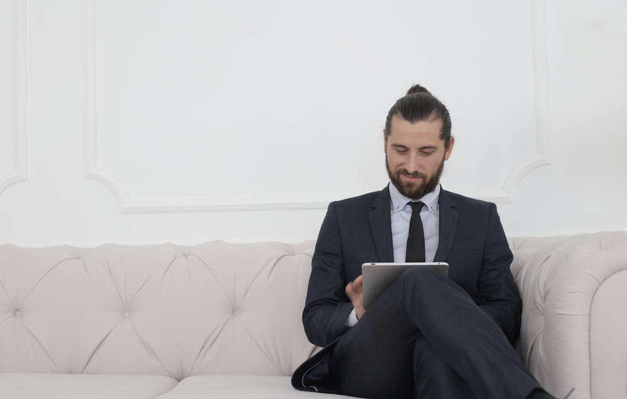 gallery-02 A businessman in a suit sitting on a sofa using a tablet, smiling.
