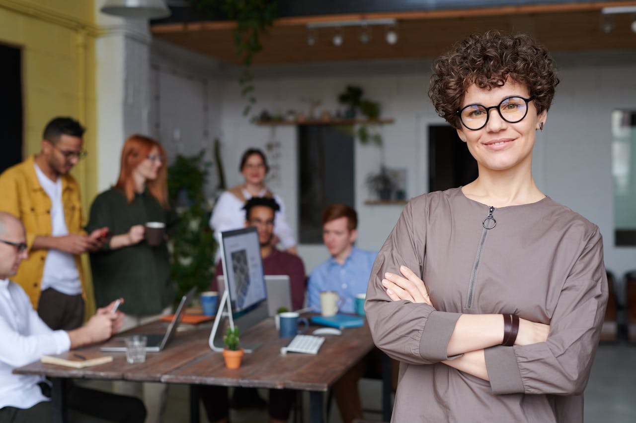 embark Smiling businesswoman with curly hair stands confidently in a modern office space with colleagues.